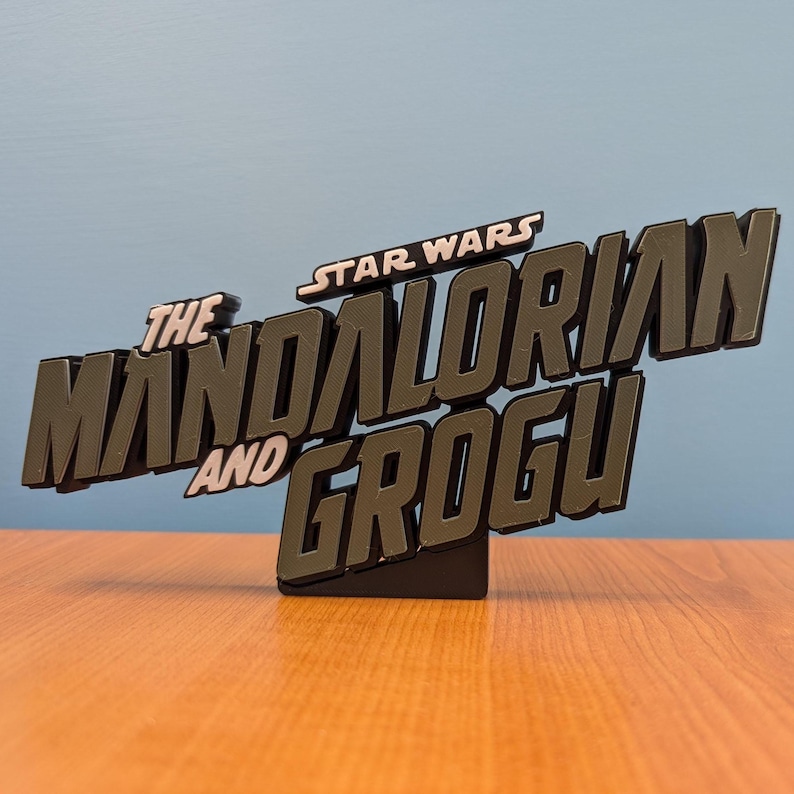 Op de afbeelding: Een zwart en grijs bord met de tekst "The Mandalorian and Grogu" en "Star Wars" erboven. Het bord is 3D en staat op een kleine zwarte basis. Een decoratief item.