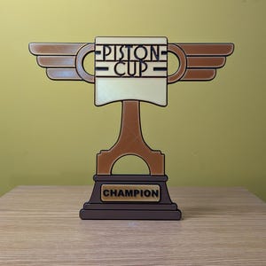 May include: A brown and gold trophy with wings, a piston base, and a white cup with the text "PISTON CUP" on it. The base of the trophy has the word "CHAMPION" on it.