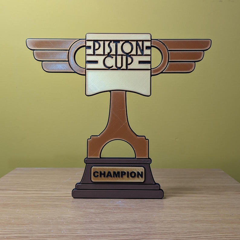 May include: A brown and gold trophy with wings, a piston base, and a white cup with the text "PISTON CUP" on it. The base of the trophy has the word "CHAMPION" on it.