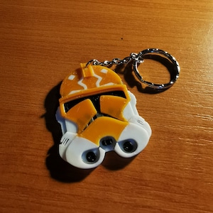 May include: A yellow and white 3D printed keychain featuring a Star Wars clone trooper helmet. The keychain has a silver metal ring.