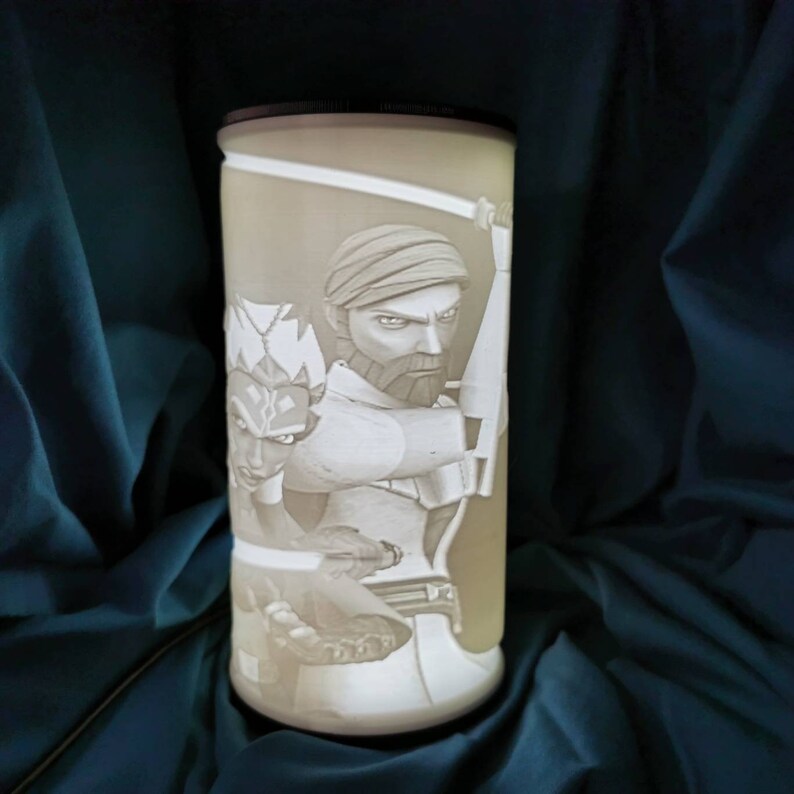 May include: A white cylindrical lamp with a 3D image of two characters from Star Wars: The Clone Wars. The characters are Ahsoka Tano and Anakin Skywalker. The image is lit from behind, creating a glowing effect.