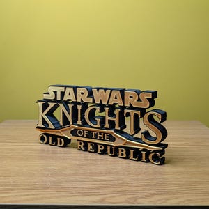 Może przedstawiać: Tr&oacute;jwymiarowy model logo gry Star Wars Knights of the Old Republic, wydrukowany w technologii 3D. Logo jest czarne z złotymi akcentami i zawiera napis "Star Wars Knights of the Old Republic".