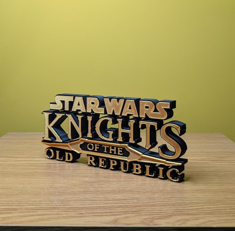 Może przedstawiać: Tr&oacute;jwymiarowy model logo gry Star Wars Knights of the Old Republic, wydrukowany w technologii 3D. Logo jest czarne z złotymi akcentami i zawiera napis "Star Wars Knights of the Old Republic".