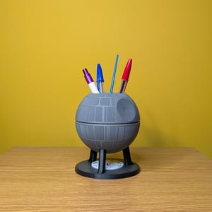 May include: A gray Death Star-shaped desk organizer with three pens inside. The pens are red, blue, and purple.