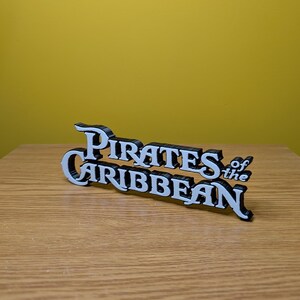 May include: White 3D printed sign with the text "Pirates of the Caribbean" in a stylized font.