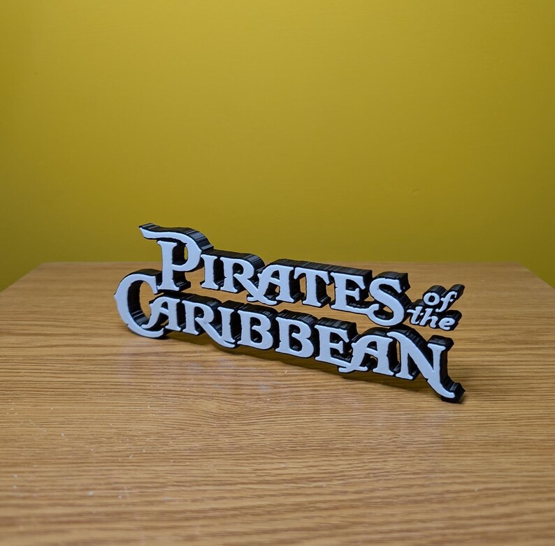 May include: White 3D printed sign with the text "Pirates of the Caribbean" in a stylized font.