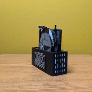 May include: A black and gray Star Wars: The Bad Batch themed coaster set with a design of a spaceship. The coasters are in a black holder with the words "Star Wars The Bad Batch" printed on the front.