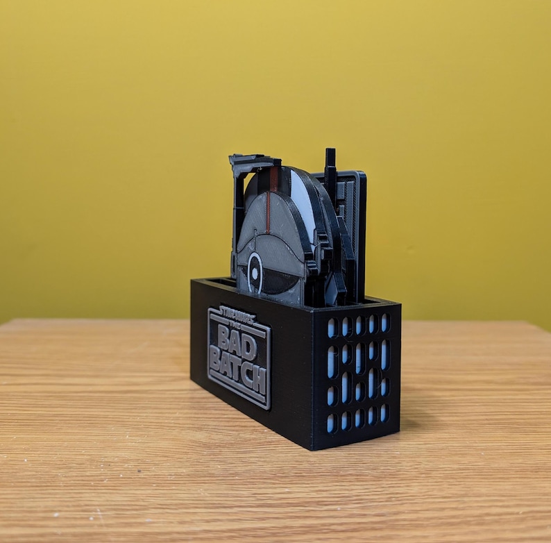 May include: A black and gray Star Wars: The Bad Batch themed coaster set with a design of a spaceship. The coasters are in a black holder with the words "Star Wars The Bad Batch" printed on the front.