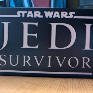 以下が含まれることがあります： 「STAR WARS JEDI SURVIVOR」と白文字で書かれた黒い看板です。