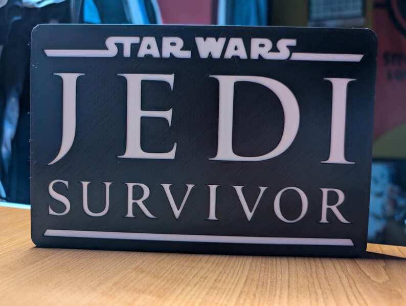 以下が含まれることがあります： 「STAR WARS JEDI SURVIVOR」と白文字で書かれた黒い看板です。