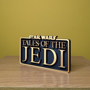 May include: A black and gold sign with the text "STAR WARS TALES OF THE JEDI". The sign is rectangular and has a gold border. The letters are gold and stand out against the black background.
