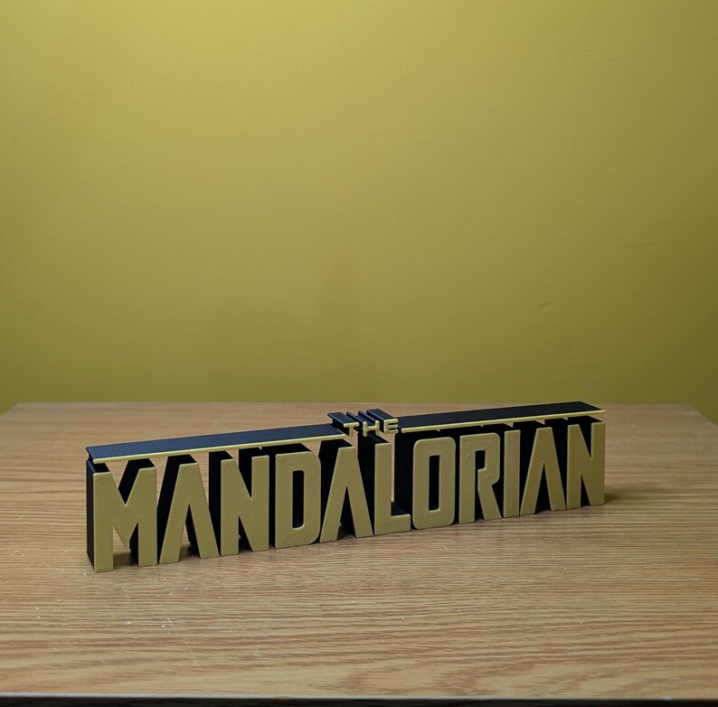 May include: A black and gold 3D printed sign that reads "THE MANDALORIAN".