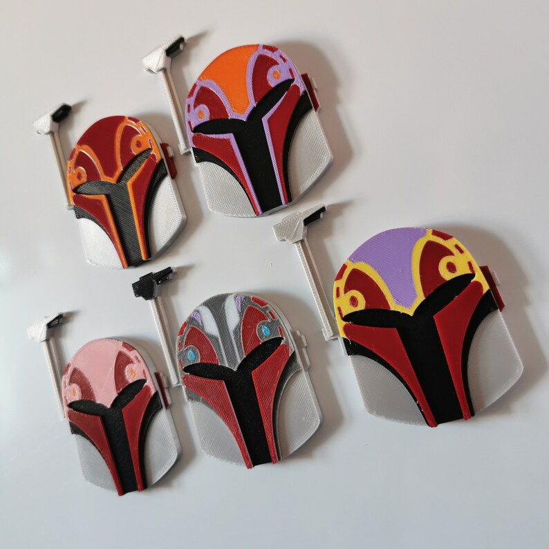 May include: Five 3D printed Mandalorian helmets in different color combinations. The helmets are red, black, white, orange, and purple with various patterns and designs.