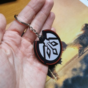 May include: A black and white keychain with a red outline. The keychain features a stylised symbol with a white background and a black design. The symbol is surrounded by a red circle.