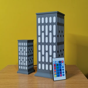 May include: Two gray, rectangular lamps with white panels and a remote control with color changing lights. The lamps are shaped like buildings.