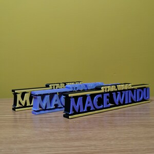 May include: Three 3D printed nameplates with the text "STAR WARS MACE WINDU" in black, gold, and purple lettering. The nameplates are stacked on top of each other.