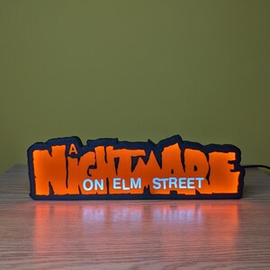 May include: A black and orange LED sign with the text "A NIGHTMARE ON ELM STREET". The sign is illuminated with orange light, and the text "ON ELM STREET" is in white. The sign is on a wooden surface.