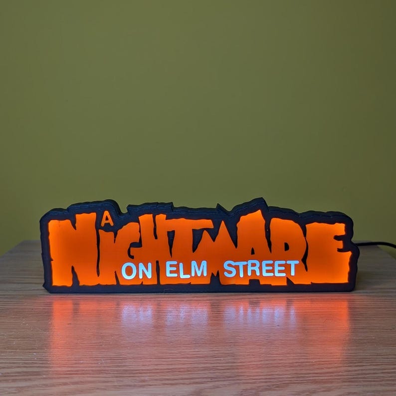 May include: A black and orange LED sign with the text "A NIGHTMARE ON ELM STREET". The sign is illuminated with orange light, and the text "ON ELM STREET" is in white. The sign is on a wooden surface.