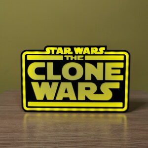 以下が含まれることがあります： 黒と黄色の看板に、太字の白いフォントで「スター・ウォーズ クローン・ウォーズ」と書かれています。