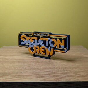 May include: A 3D printed sign with the words "Star Wars Skeleton Crew" in orange and silver letters on a black background.