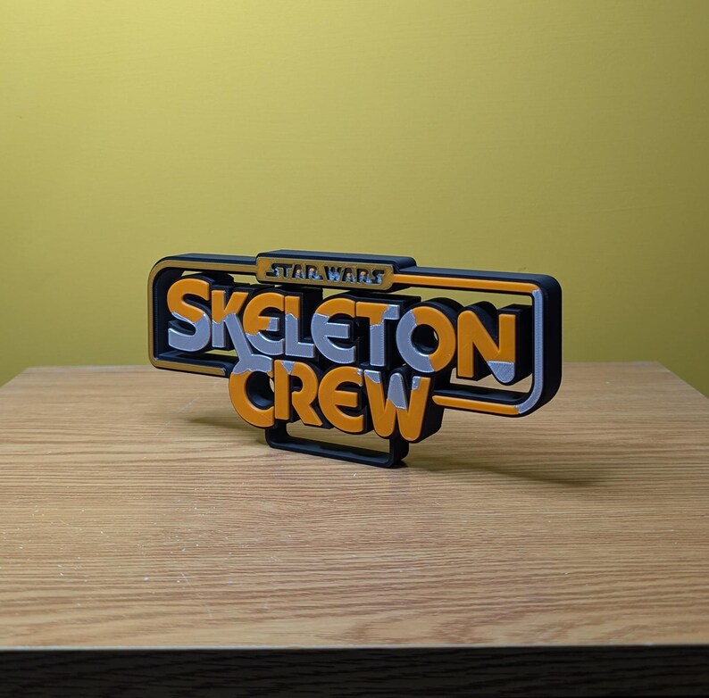 May include: A 3D printed sign with the words "Star Wars Skeleton Crew" in orange and silver letters on a black background.