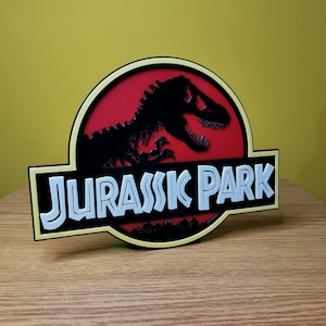 May include: A yellow and black 3D printed sign with the words "Jurassic Park" in white letters. The sign features a red background with a black silhouette of a dinosaur.