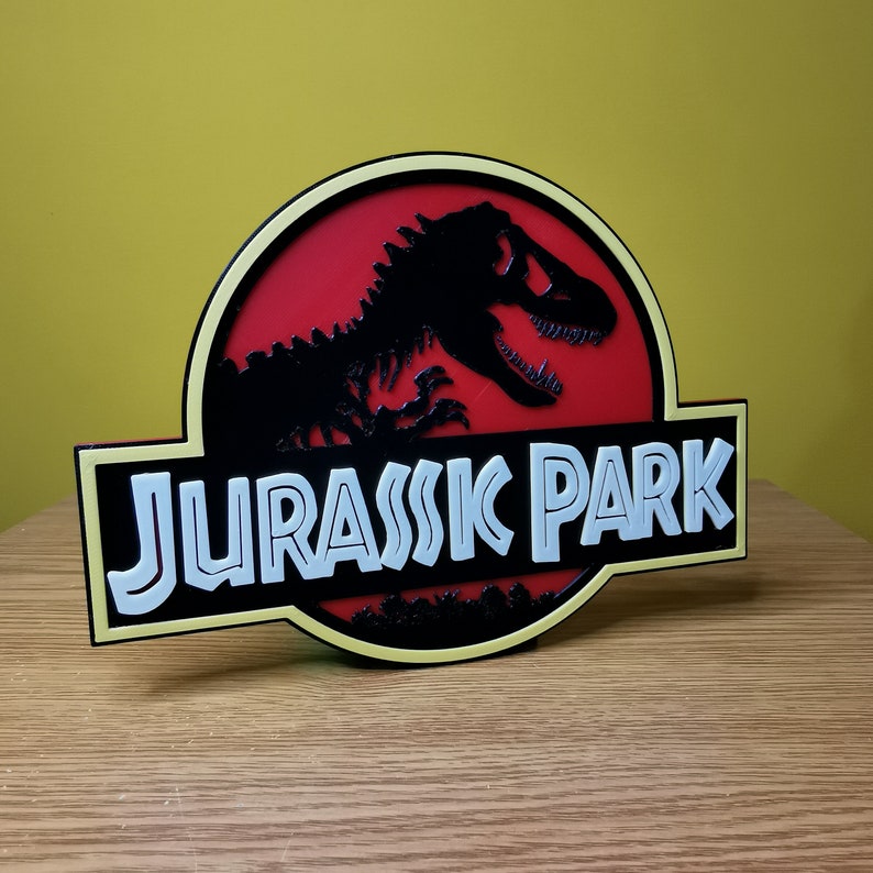 May include: A yellow and black 3D printed sign with the words "Jurassic Park" in white letters. The sign features a red background with a black silhouette of a dinosaur.