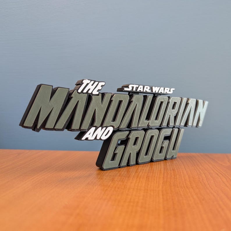 Op de afbeelding: Een 3D-geprint bord met de tekst "THE MANDALORIAN AND GROGU!". De woorden zijn in wit, zwart en olijfgroen. Het Star Wars-logo staat boven het woord "Mandalorian".