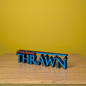 May include: A black and blue 3D printed sign that reads "STAR WARS THRAWN". The sign is on a wooden surface.
