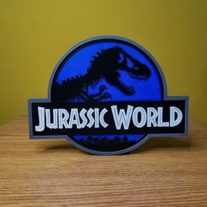 May include: A blue and gray 3D printed sign with the Jurassic World logo. The logo features a black silhouette of a dinosaur with a white outline. The text "Jurassic World" is in white with a black outline.