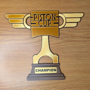 May include: A gold-colored trophy with a piston-shaped base and wings. The trophy has the words "PISTONE CUP" on the front and "CHAMPION" on the base.