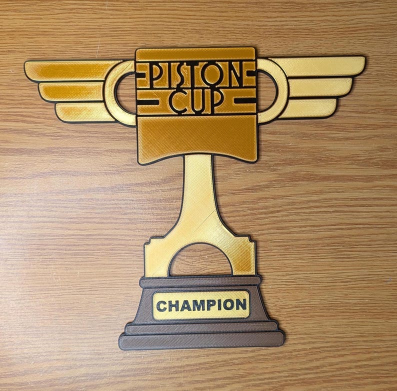 May include: A gold-colored trophy with a piston-shaped base and wings. The trophy has the words "PISTONE CUP" on the front and "CHAMPION" on the base.