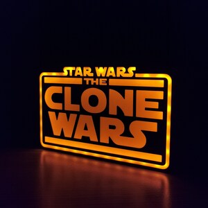 以下が含まれることがあります： 黒い長方形の枠の中に「STAR WARS THE CLONE WARS」という文字が書かれた、黄色く光る看板です。