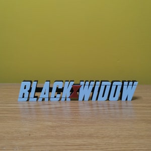 May include: A white 3D printed sign with the words "BLACK WIDOW" in black lettering. The "W" in Widow is red.