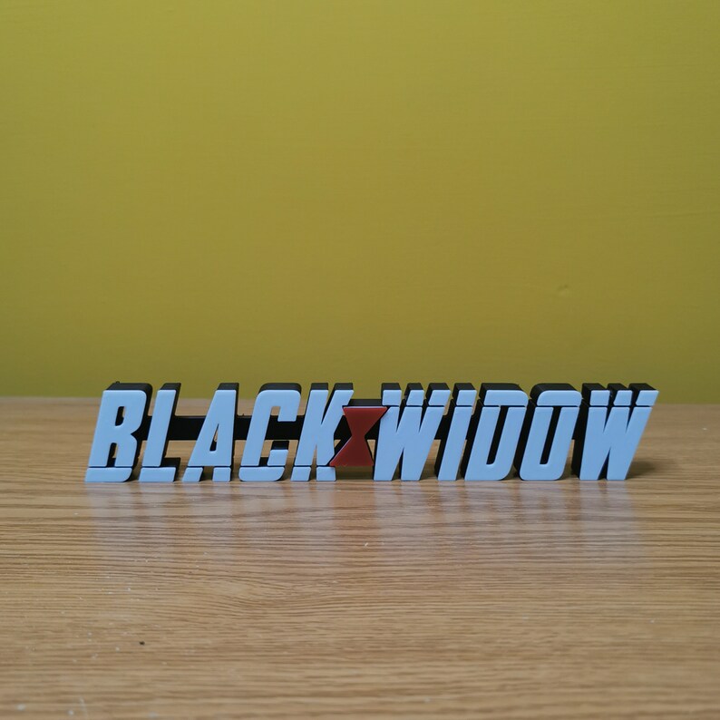 May include: A white 3D printed sign with the words "BLACK WIDOW" in black lettering. The "W" in Widow is red.