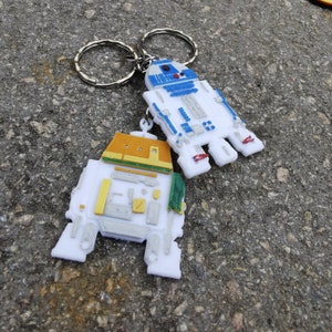 May include: Two white robot keychains with silver key rings. One robot is blue and white, the other is yellow and white.