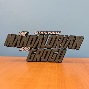 Op de afbeelding: Een donkergrijs, driedimensionaal bord met de tekst "THE MANDALORIAN AND GROGU" en "STAR WARS" erboven. Het bord staat tegen een lichtblauwe achtergrond en rust op een houten oppervlak.