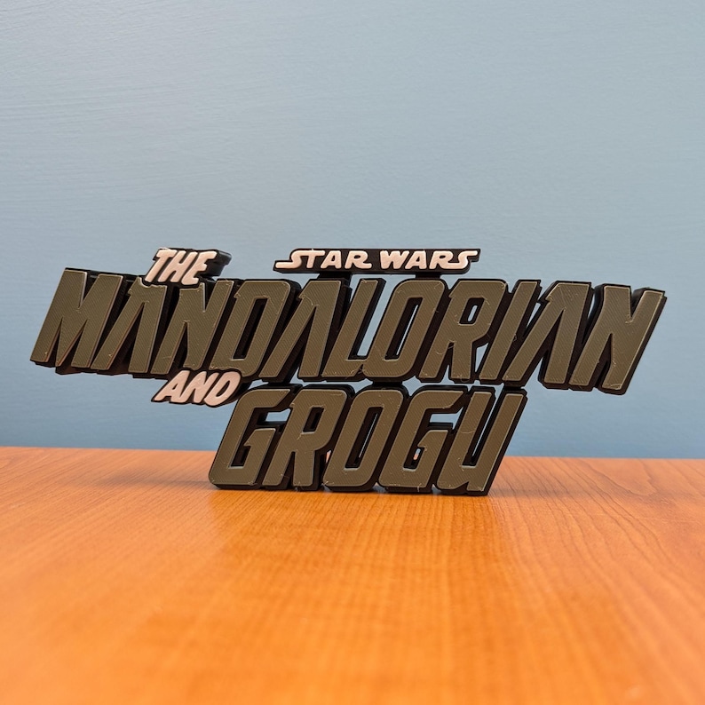 Op de afbeelding: Een donkergrijs, driedimensionaal bord met de tekst "THE MANDALORIAN AND GROGU" en "STAR WARS" erboven. Het bord staat tegen een lichtblauwe achtergrond en rust op een houten oppervlak.