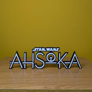 May include: A white 3D printed sign with the text "STAR WARS AHSOKA" in black outlined letters. The sign is on a wooden surface.