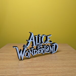 Pode incluir: Uma placa preta e branca que diz "Alice in Wonderland" em uma fonte estilizada. A placa &eacute; colocada em uma superf&iacute;cie de madeira com um fundo amarelo.