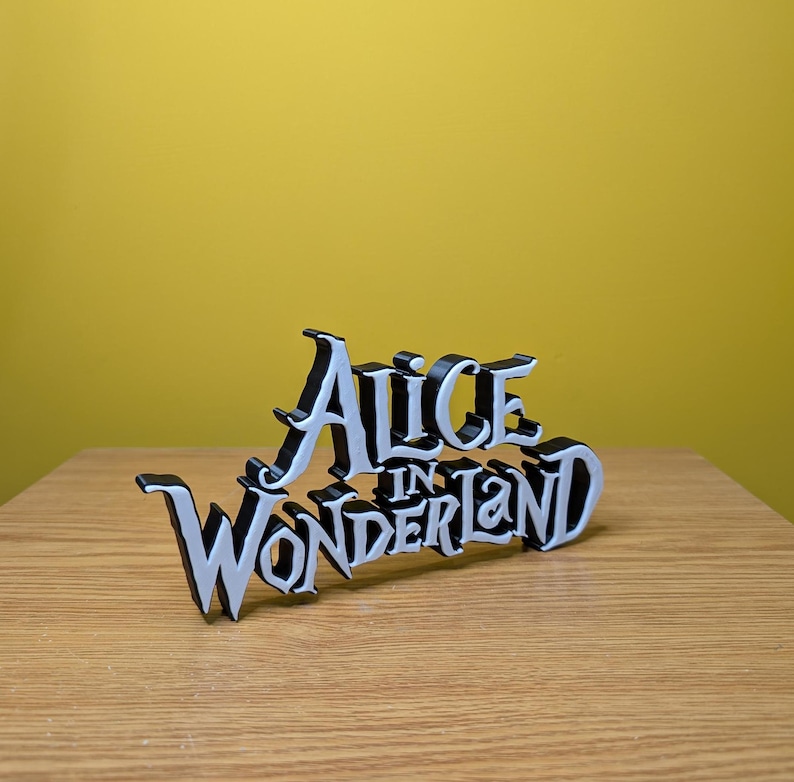 Pode incluir: Uma placa preta e branca que diz "Alice in Wonderland" em uma fonte estilizada. A placa &eacute; colocada em uma superf&iacute;cie de madeira com um fundo amarelo.