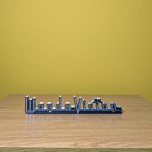 Peut inclure: Logo "WandaVision" argent&eacute; et noir pos&eacute; sur une surface en bois devant un fond jaune. Le logo est une conception tridimensionnelle, avec le texte en gras, une police sans empattement. Les lettres sont soulign&eacute;es en noir, avec un remplissage argent&eacute;.
