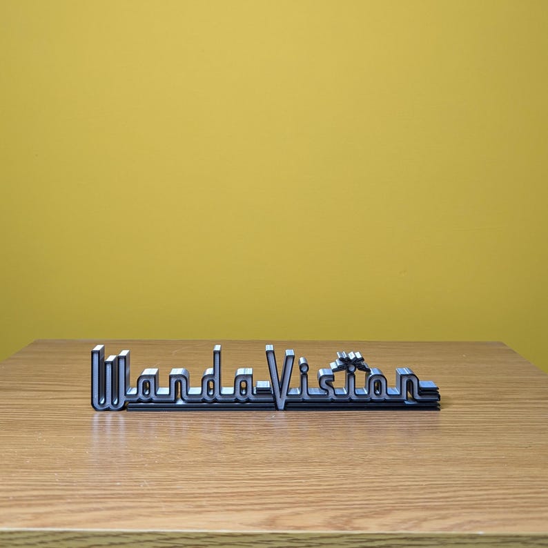 Peut inclure: Logo "WandaVision" argent&eacute; et noir pos&eacute; sur une surface en bois devant un fond jaune. Le logo est une conception tridimensionnelle, avec le texte en gras, une police sans empattement. Les lettres sont soulign&eacute;es en noir, avec un remplissage argent&eacute;.