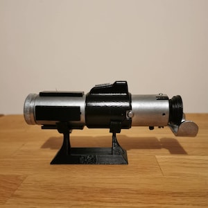 K&ouml;nnte beinhalten: Ein silberner und schwarzer Lichtschwert-Prop auf einem schwarzen Plastikst&auml;nder. Das Lichtschwert hat einen schwarzen Griff und eine silberne Klinge. Der St&auml;nder hat den Text "STAR WARS" aufgedruckt.