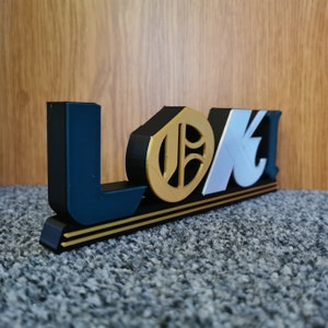 May include: A 3D printed sign with the word "LOKI" in black, white, and gold letters. The letters are raised and have a textured surface. The sign is on a black base with two gold stripes.