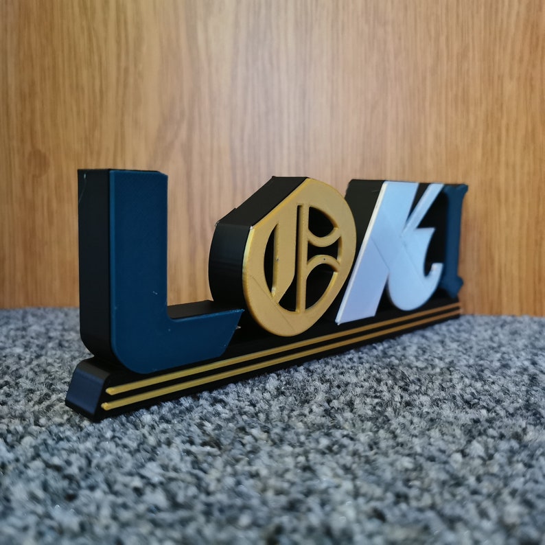 May include: A 3D printed sign with the word "LOKI" in black, white, and gold letters. The letters are raised and have a textured surface. The sign is on a black base with two gold stripes.