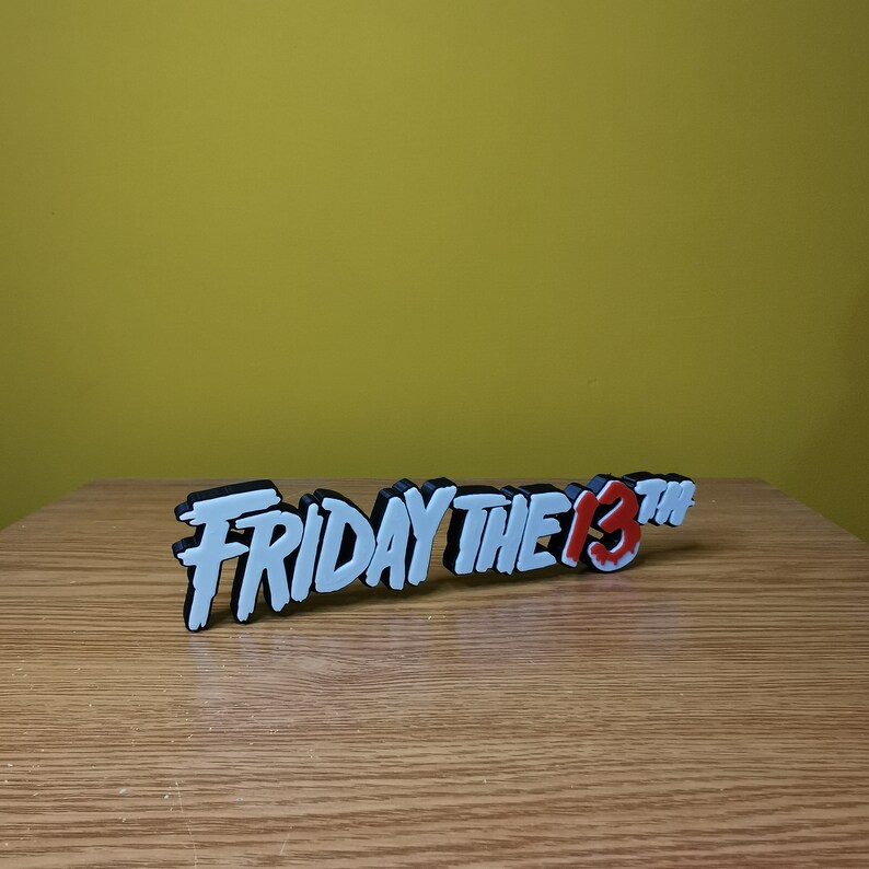 Puede incluir: Un letrero impreso en 3D en blanco y negro que dice "Viernes 13" con el "13" en rojo. El letrero est&aacute; sobre una superficie de madera.