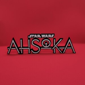 May include: A black and white "Star Wars Ahsoka" logo against a red background. The logo features the "Star Wars" text above the name "Ahsoka" in a stylized font. The letters are black with white outlines and details.