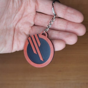 May include: A red and black round keyring with a symbol that resembles a stylised letter E. The keyring is attached to a silver chain.