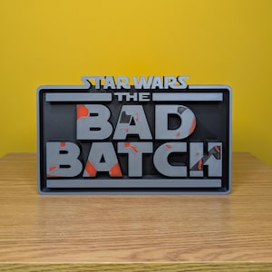 May include: A gray 3D printed sign with the text "STAR WARS THE BAD BATCH" in a distressed style. The letters are raised and have a black background with orange accents.
